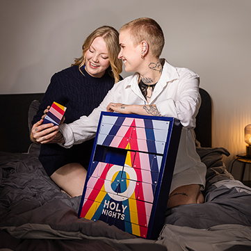 Zwei Frauen sitzen auf einem Bett und lachen, während sie eine Schublade des ORION Adventkalenders öffnen. Die Stimmung ist fröhlich und intim.